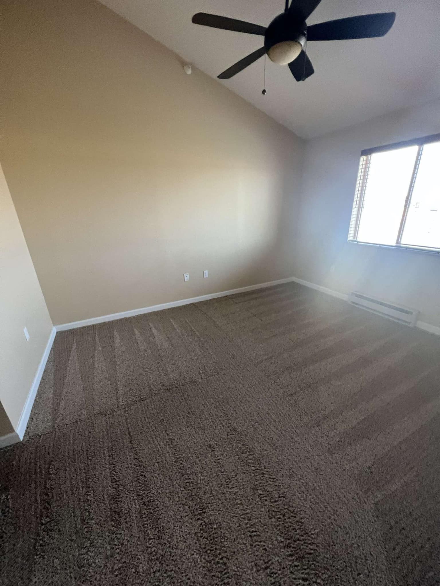 Empty room with tan walls, brown carpet, ceiling fan, and window.