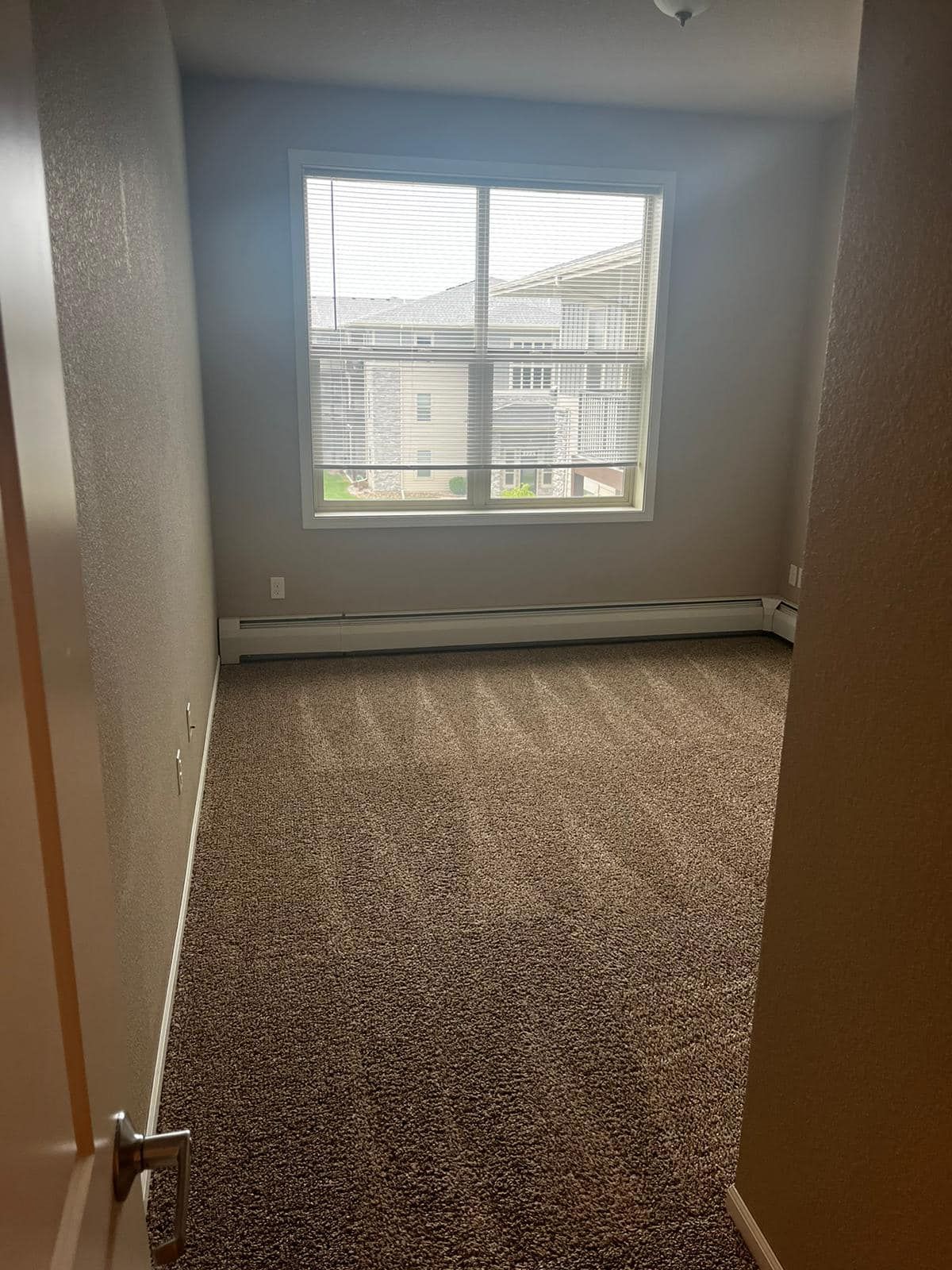 Empty bedroom with brown carpet, a window with blinds, and beige walls.
