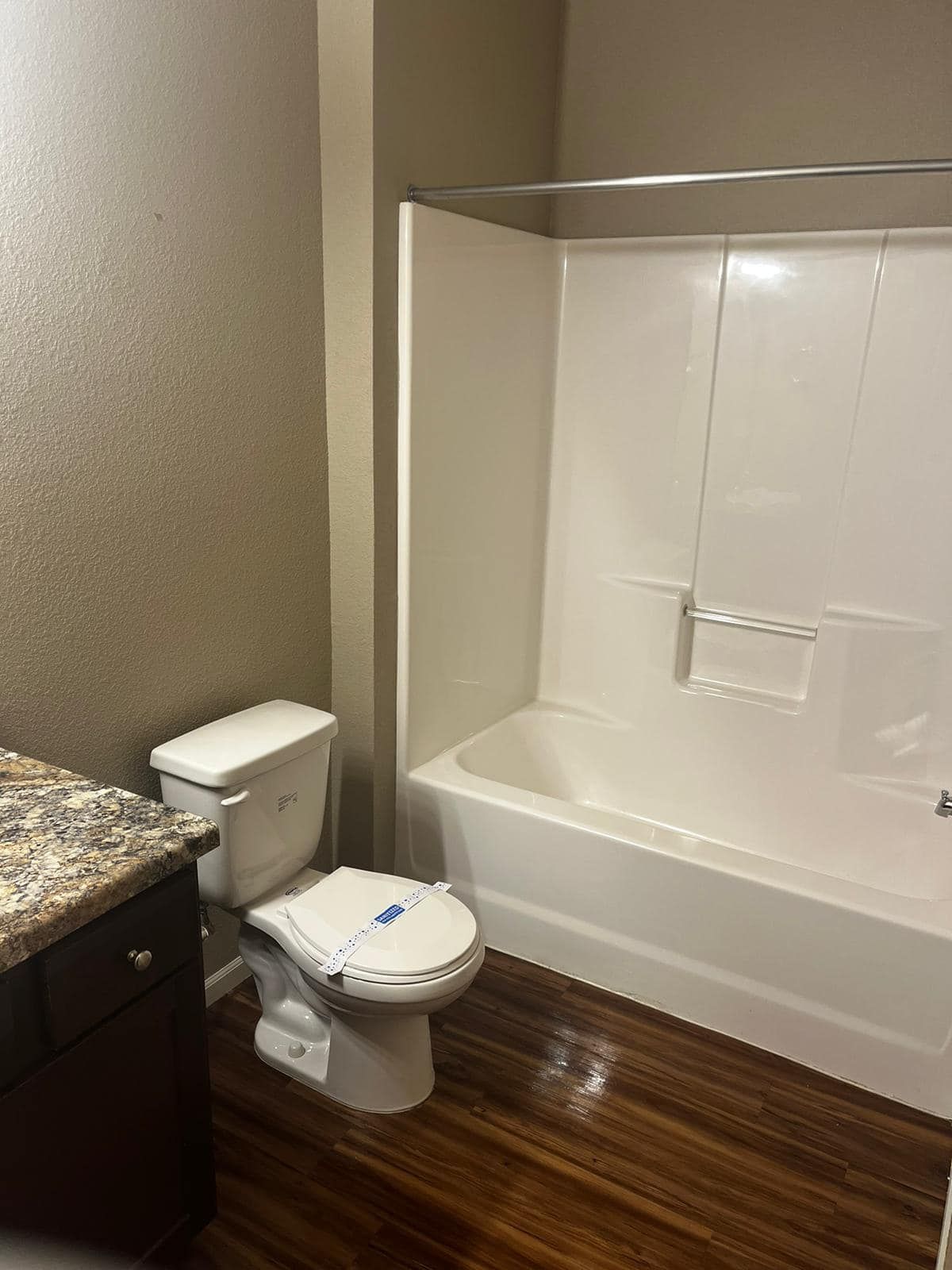 Bathroom with a toilet, shower/tub combo, and dark wood-look flooring. Brown walls.