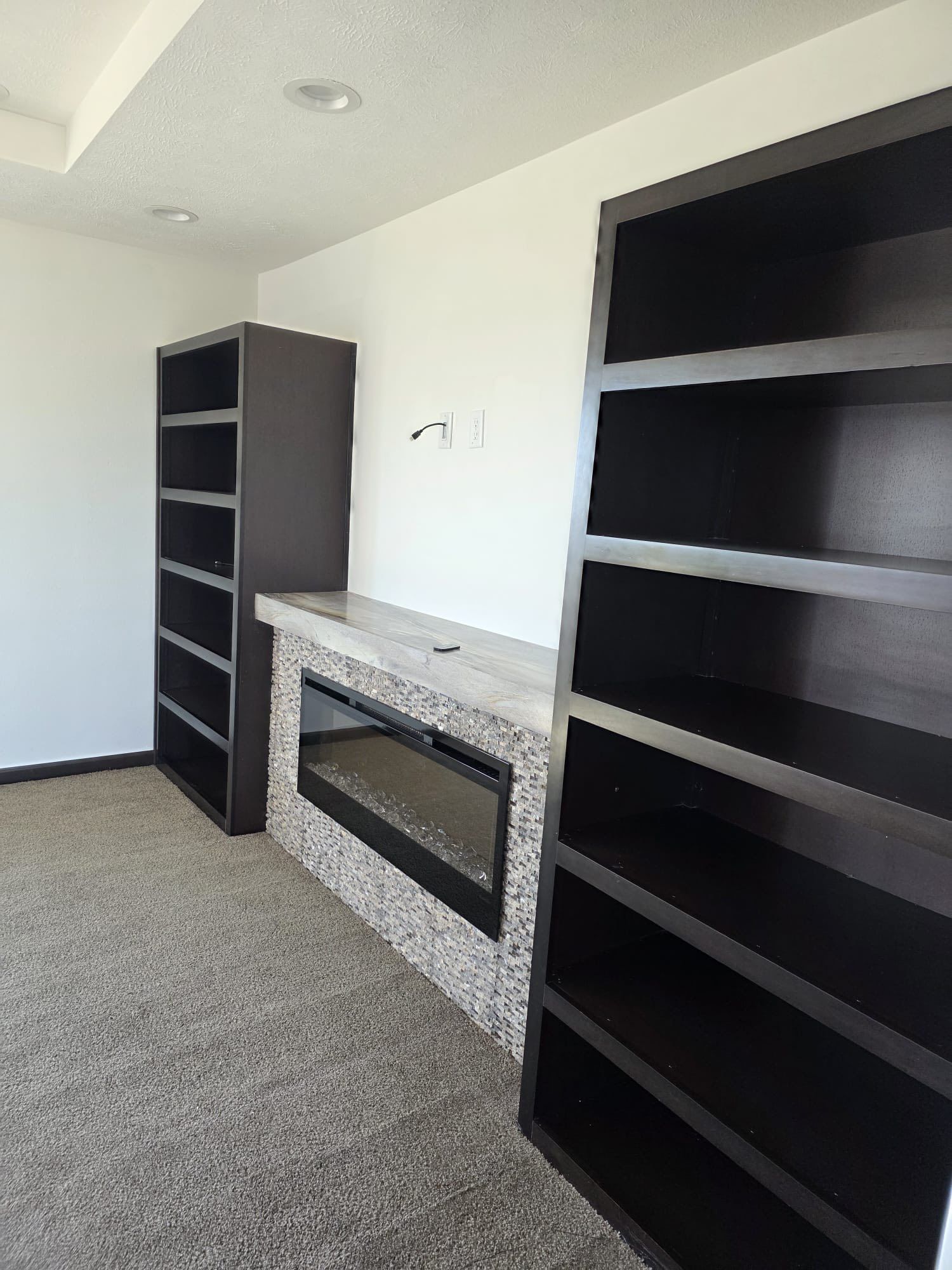 Built-in dark bookcases flanking a stone-faced electric fireplace. Room has neutral carpet and white walls.