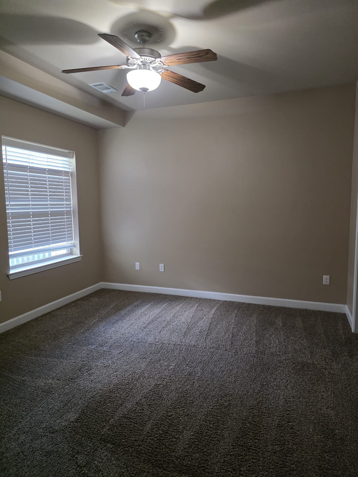 Empty bedroom with brown carpet, tan walls, ceiling fan, and window with blinds.