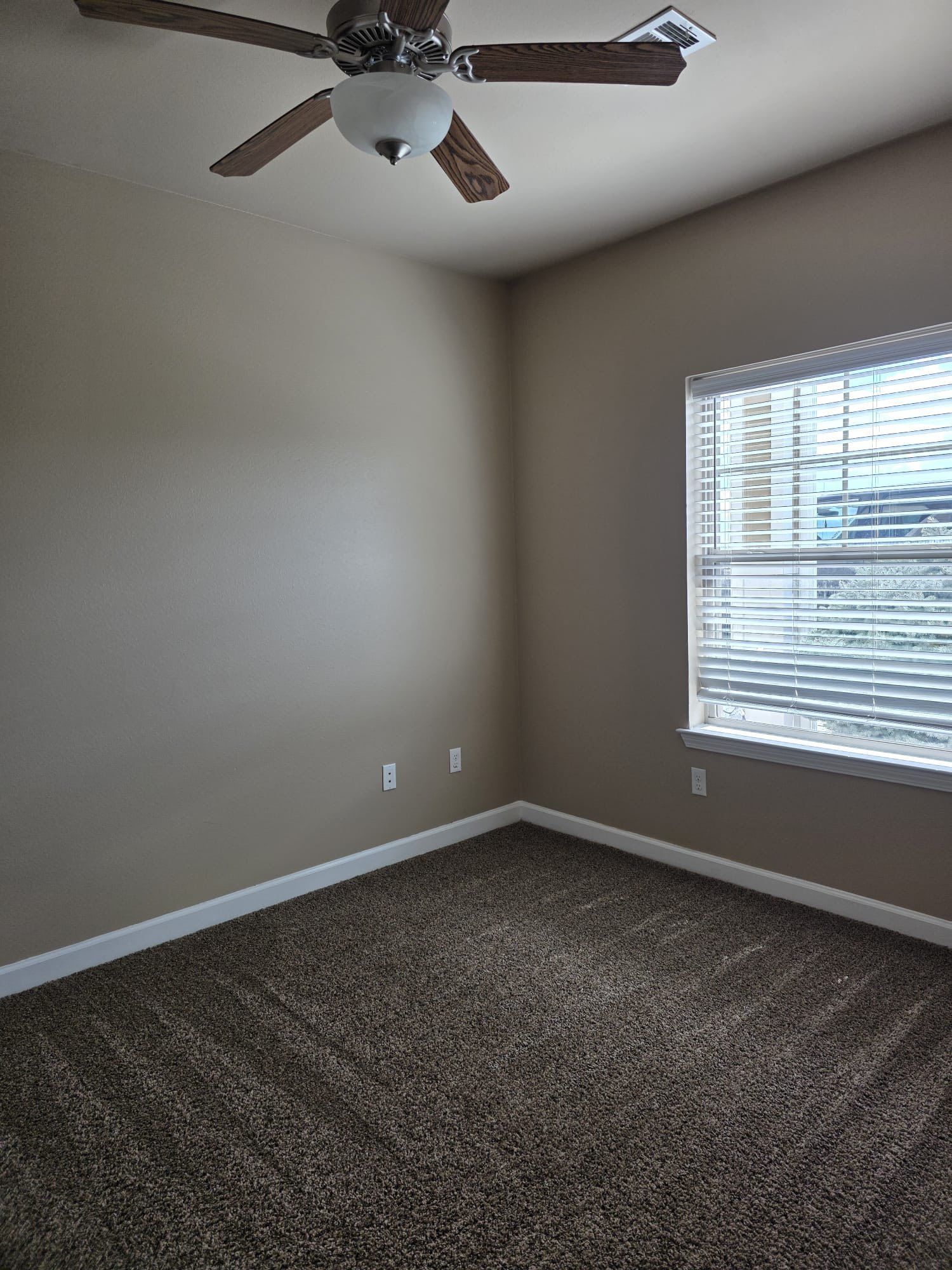 Empty room with tan walls, brown carpet, window, and ceiling fan.