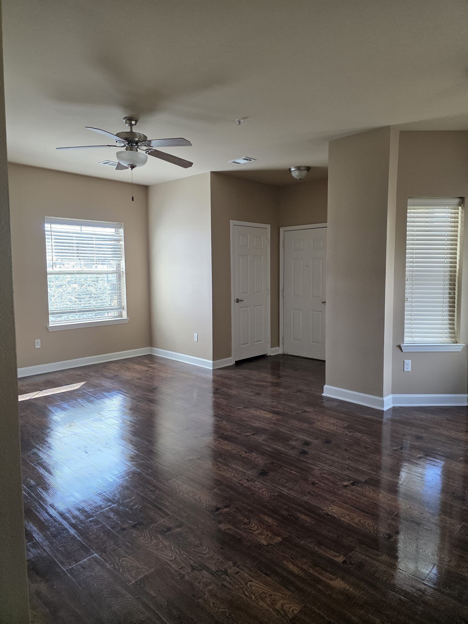 Empty living room with dark wood floors, light walls, two doors, and two windows with blinds.