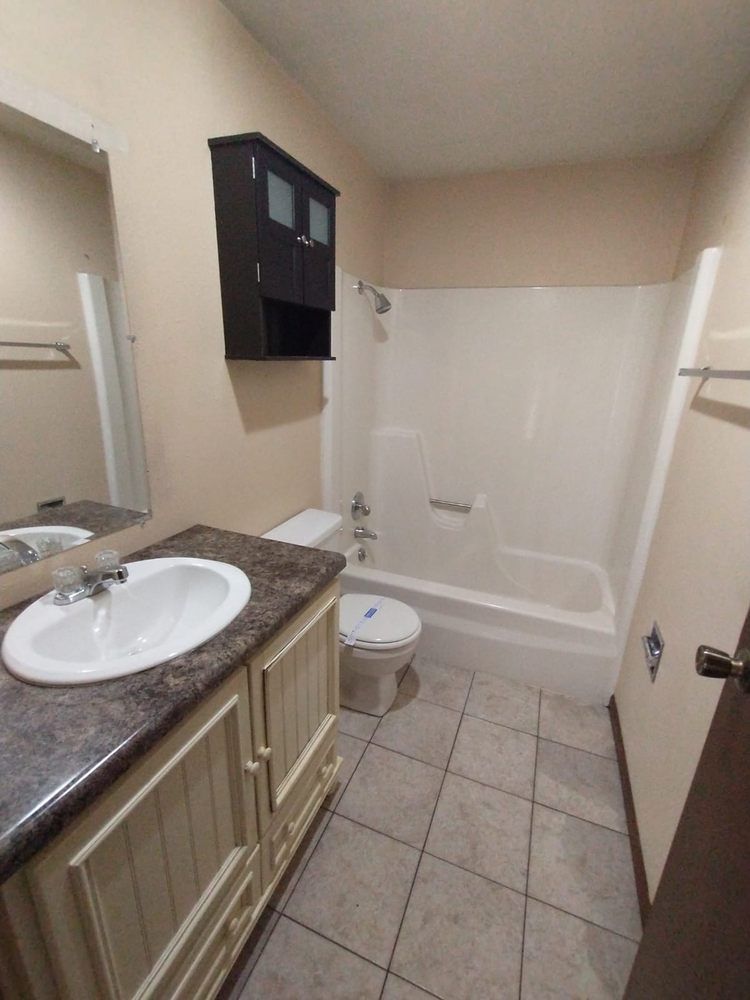 Bathroom with white tub/shower, vanity, and toilet. Beige walls, brown counter, and dark cabinet.