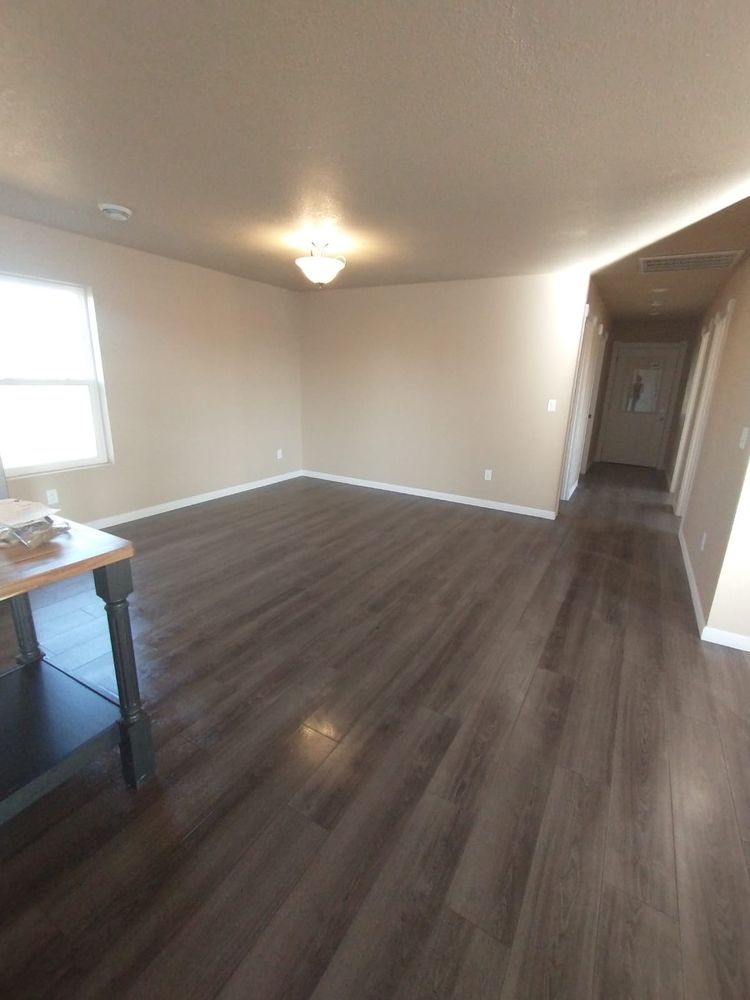 Empty living room with dark wood floors, tan walls, and a hallway.