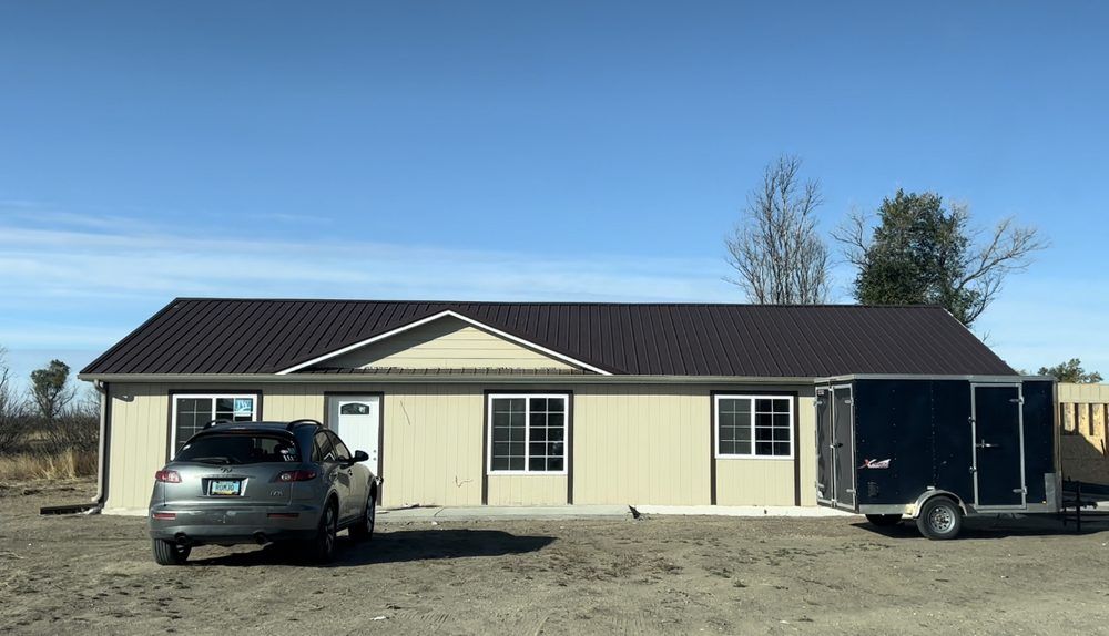 Beige building with a dark roof and white trim. A car and trailer are parked in front.