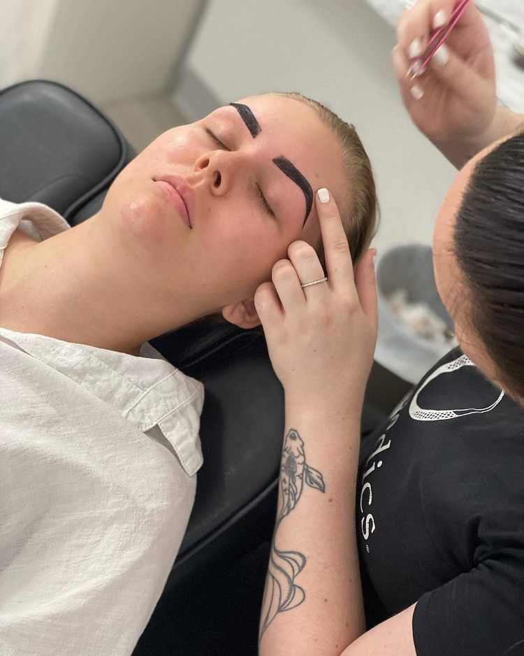 A Woman With A Tattoo On Her Arm Is Working On A Client's Brows — Bloom Skin Brows & Beauty In Bungalow, QLD