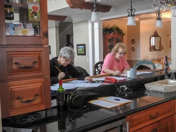 Two older women are sitting at a kitchen counter writing on papers .
