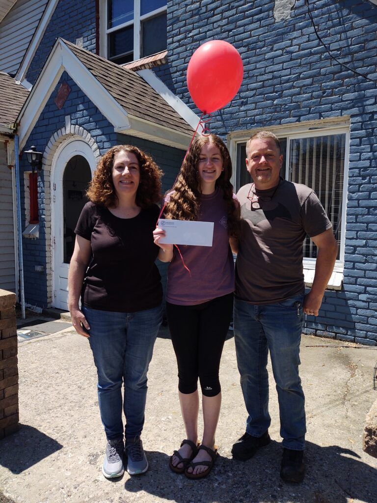 Three people standing in front of a blue brick building holding a check and a red balloon.