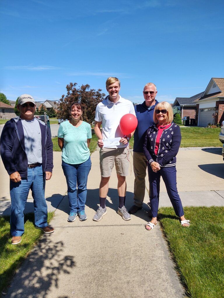 A group of people standing on a sidewalk with a man holding a red balloon and a check.