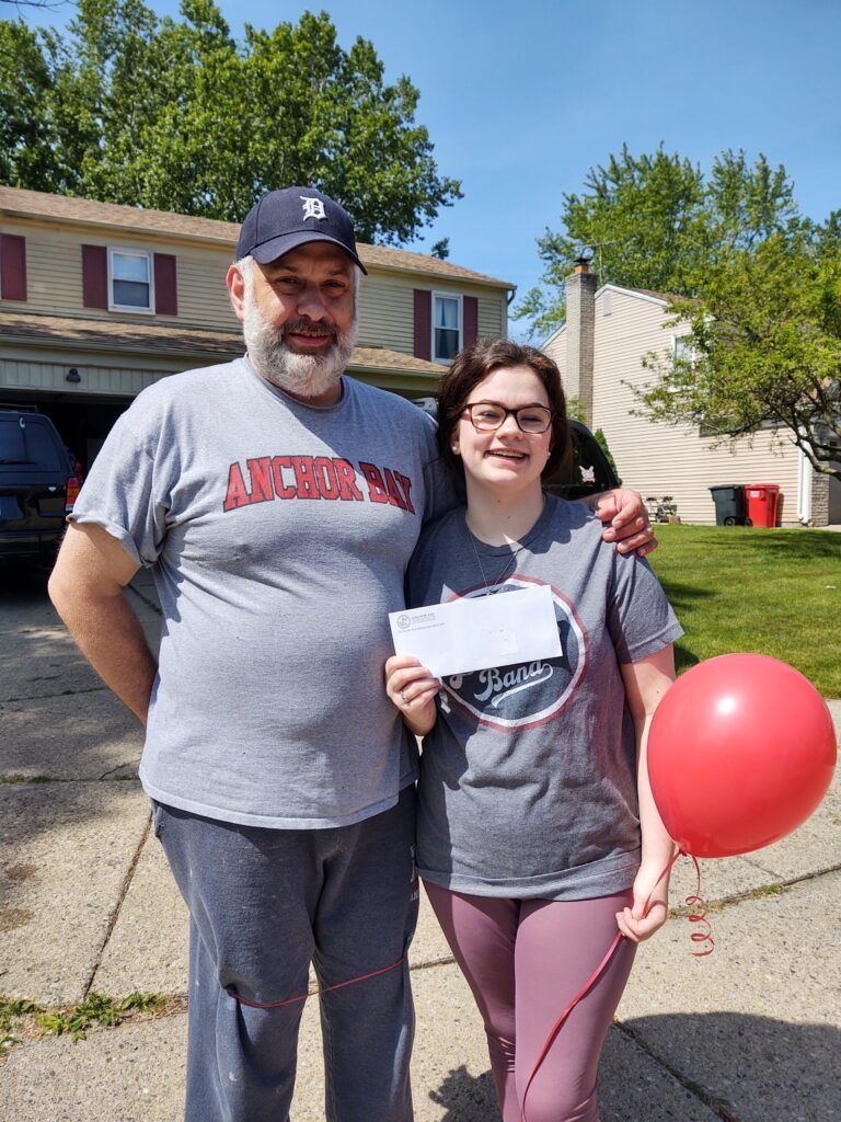 A man in an Anchor Bay shirt stands next to a girl with a red balloon and envelope.