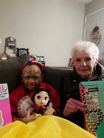 A little girl with face paint and a doll is sitting on a couch next to an older woman.