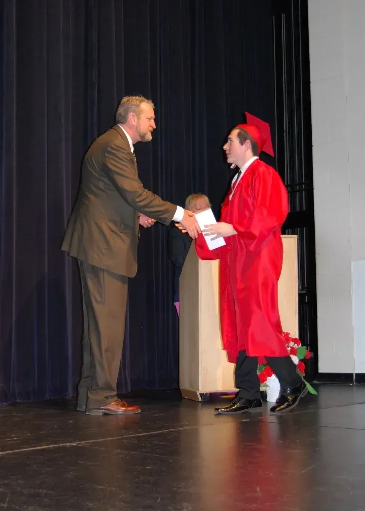 A man in a suit shakes hands with a man in a red cap and gown.