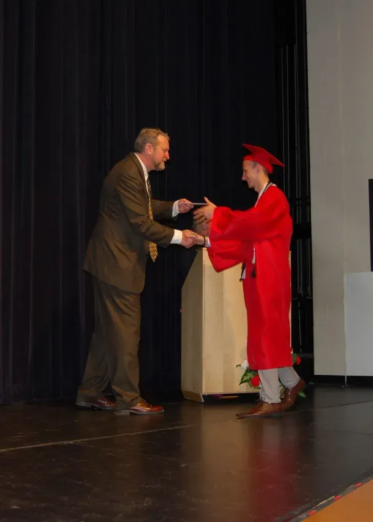 A man in a suit shakes hands with a graduate in a red cap and gown.
