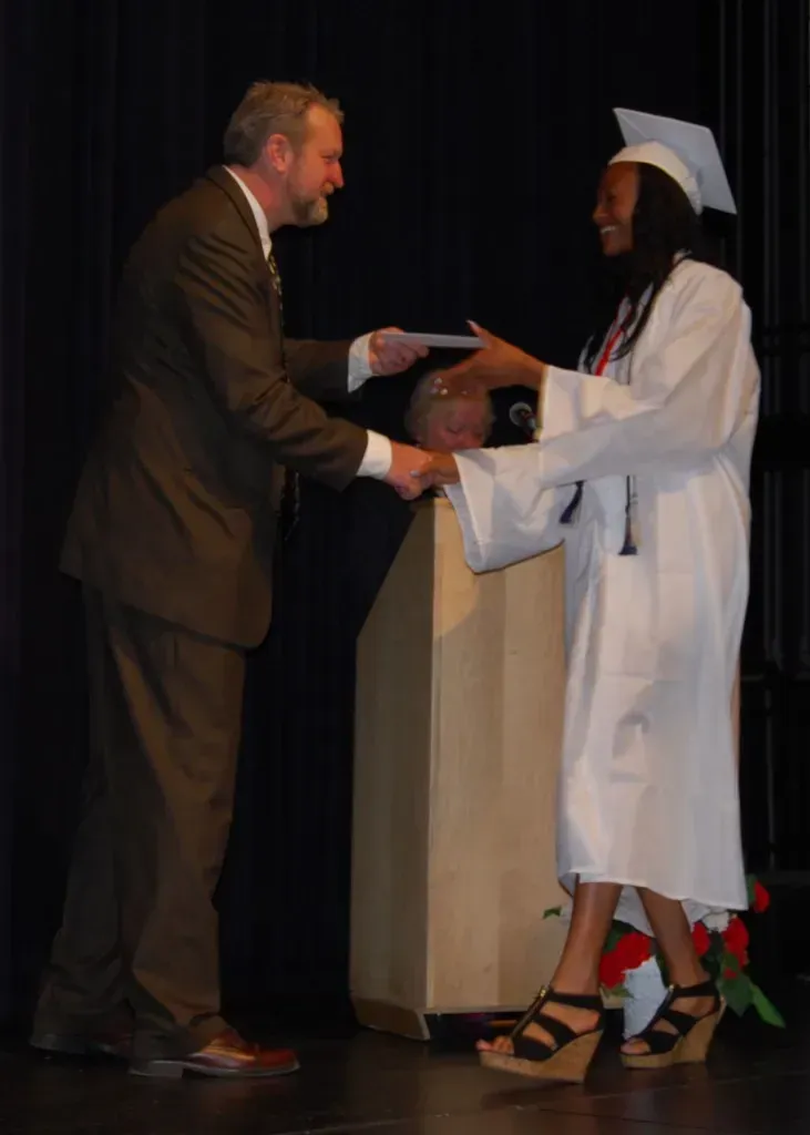 A woman in a graduation cap and gown shakes hands with a man in a suit.