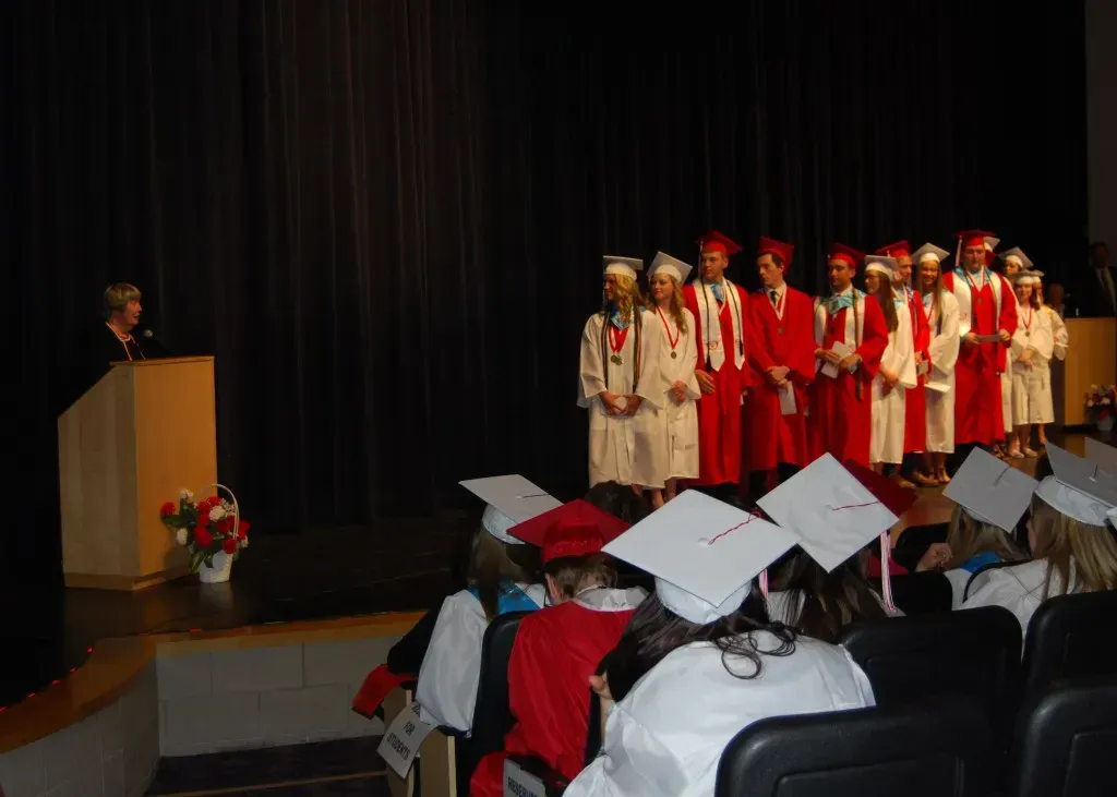 A group of graduates are standing in front of a podium while other graduates watch.