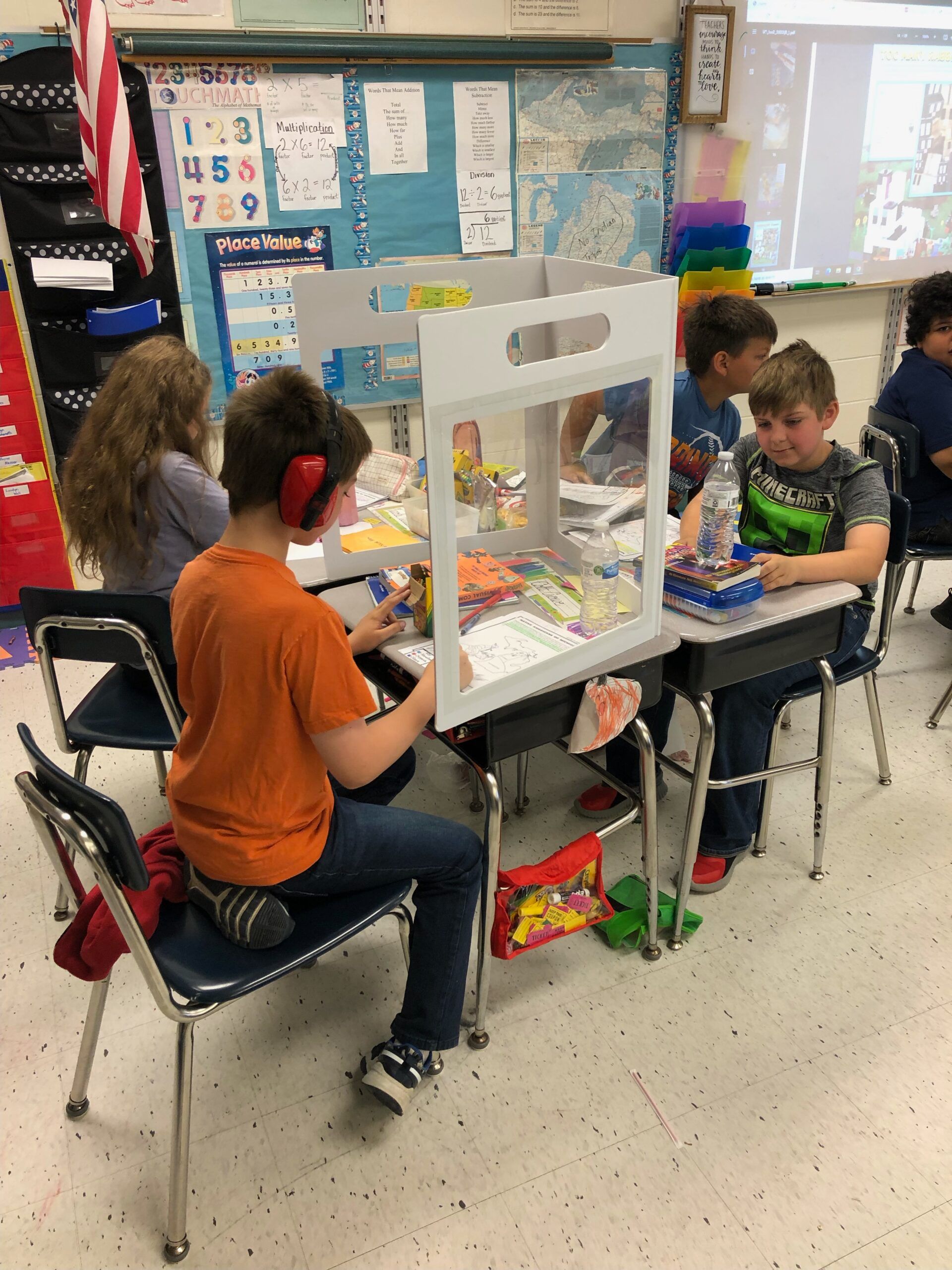 A group of children sit at desks in front of a place value chart.