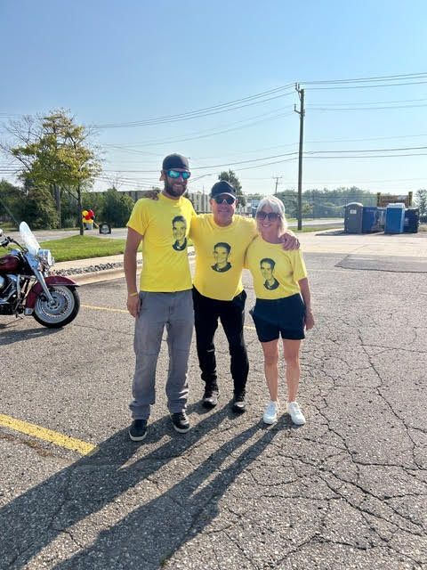 Three people in yellow t-shirts stand outside on a sunny day. A motorcycle is in the background.