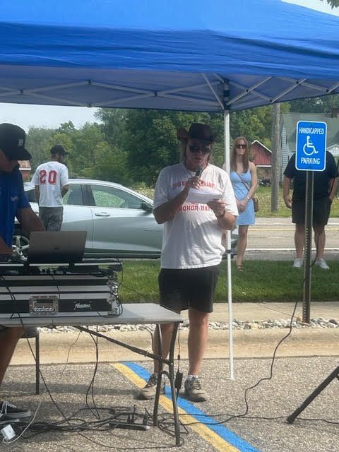 Man speaking at an outdoor event with a DJ setup, under a canopy. People watch nearby.