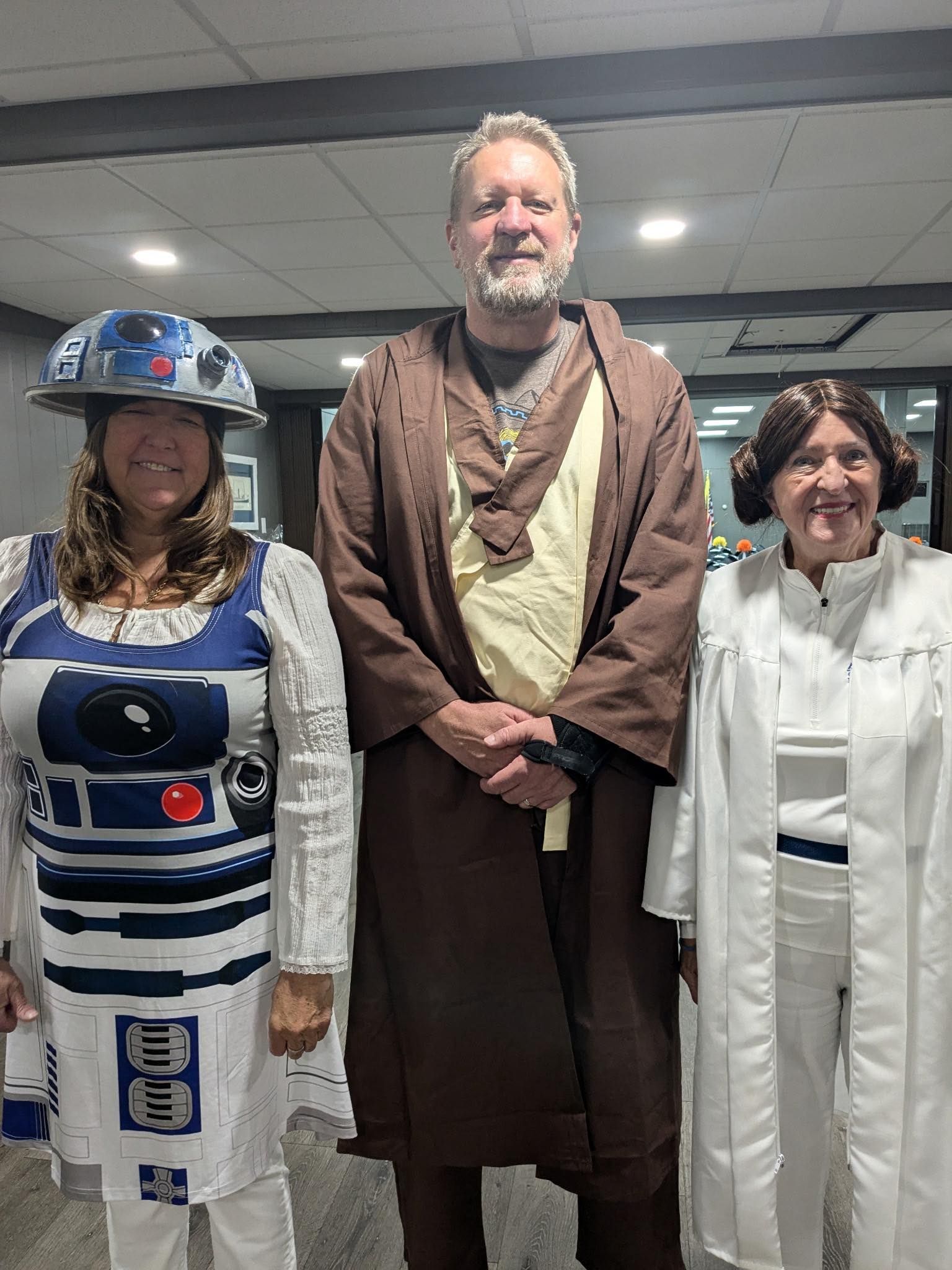 Three people in Star Wars costumes: R2-D2, Obi-Wan Kenobi, and Princess Leia pose indoors.