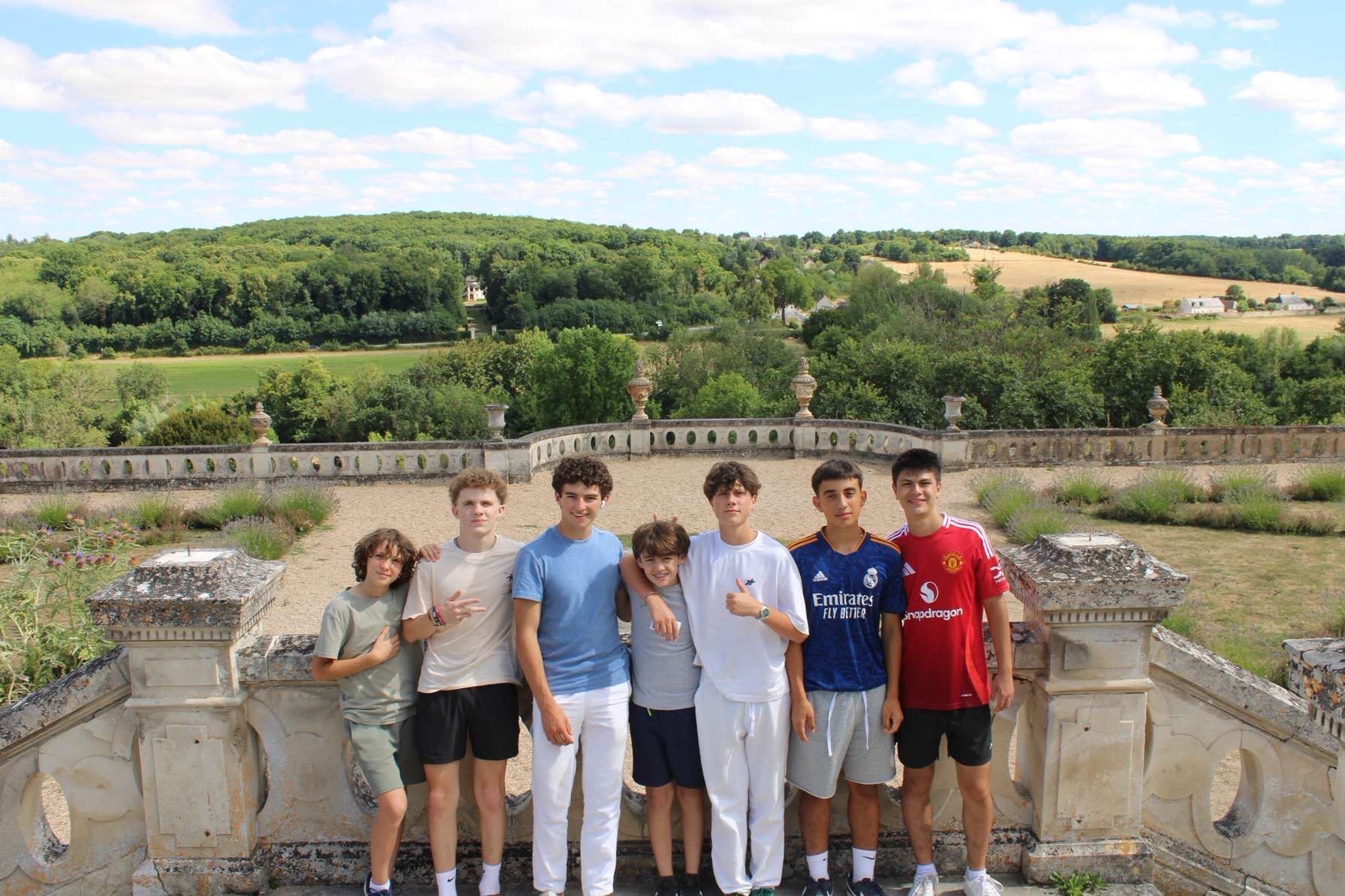 Seven young people standing on a stone structure, overlooking a green landscape under a blue sky.