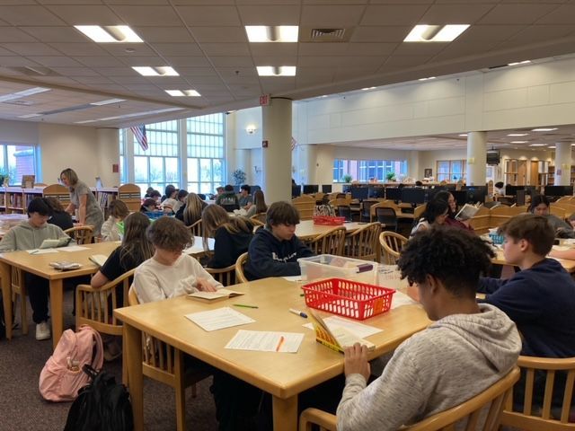 A group of students are sitting at tables in a library reading books.