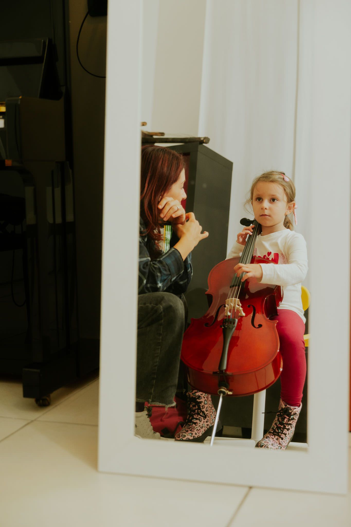 Un bambino con in mano un violoncello è seduto all'interno di una cornice bianca, con una donna accanto a lui, in un ambiente interno.