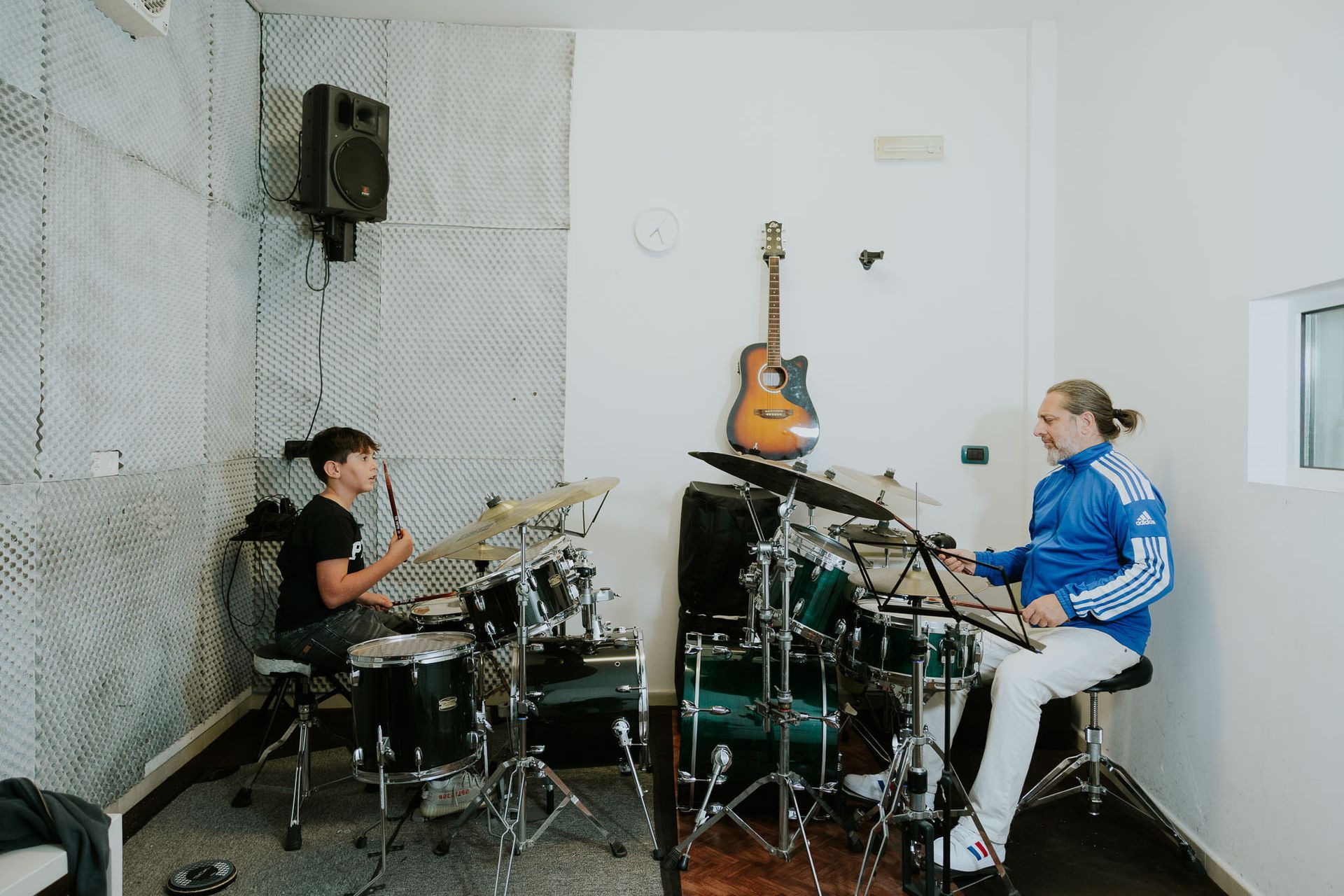 Due persone suonano la batteria in uno studio musicale; uno è un bambino. Una chitarra è appesa al muro.