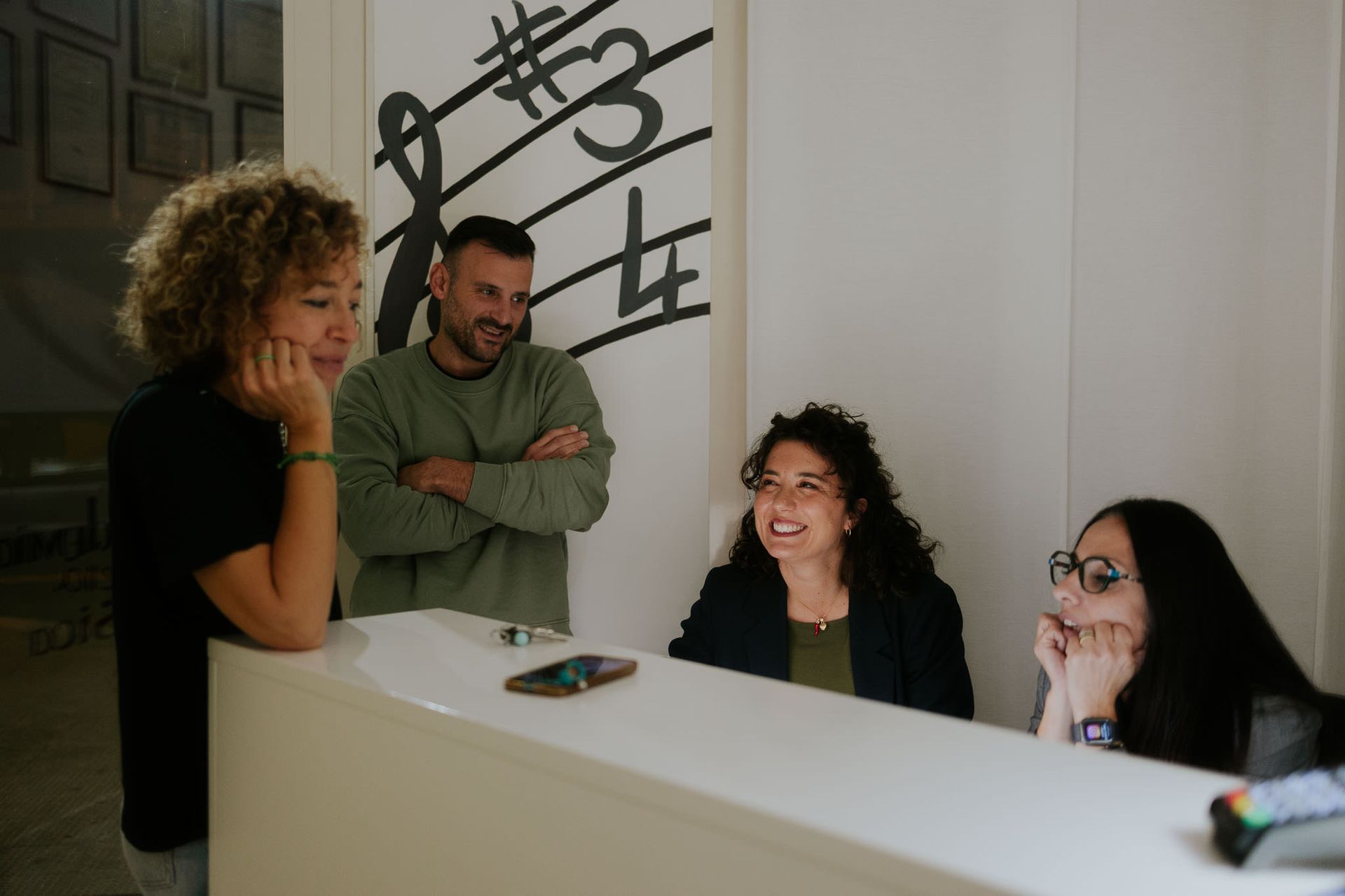 Persone sedute a un bancone bianco della reception con simboli musicali sullo sfondo; una persona è appoggiata al bancone, le altre sorridono.