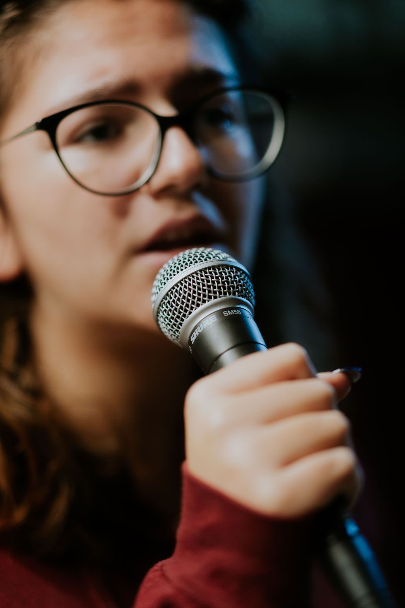 Donna che canta in un microfono, indossa occhiali e una maglietta rossa, sfondo scuro.