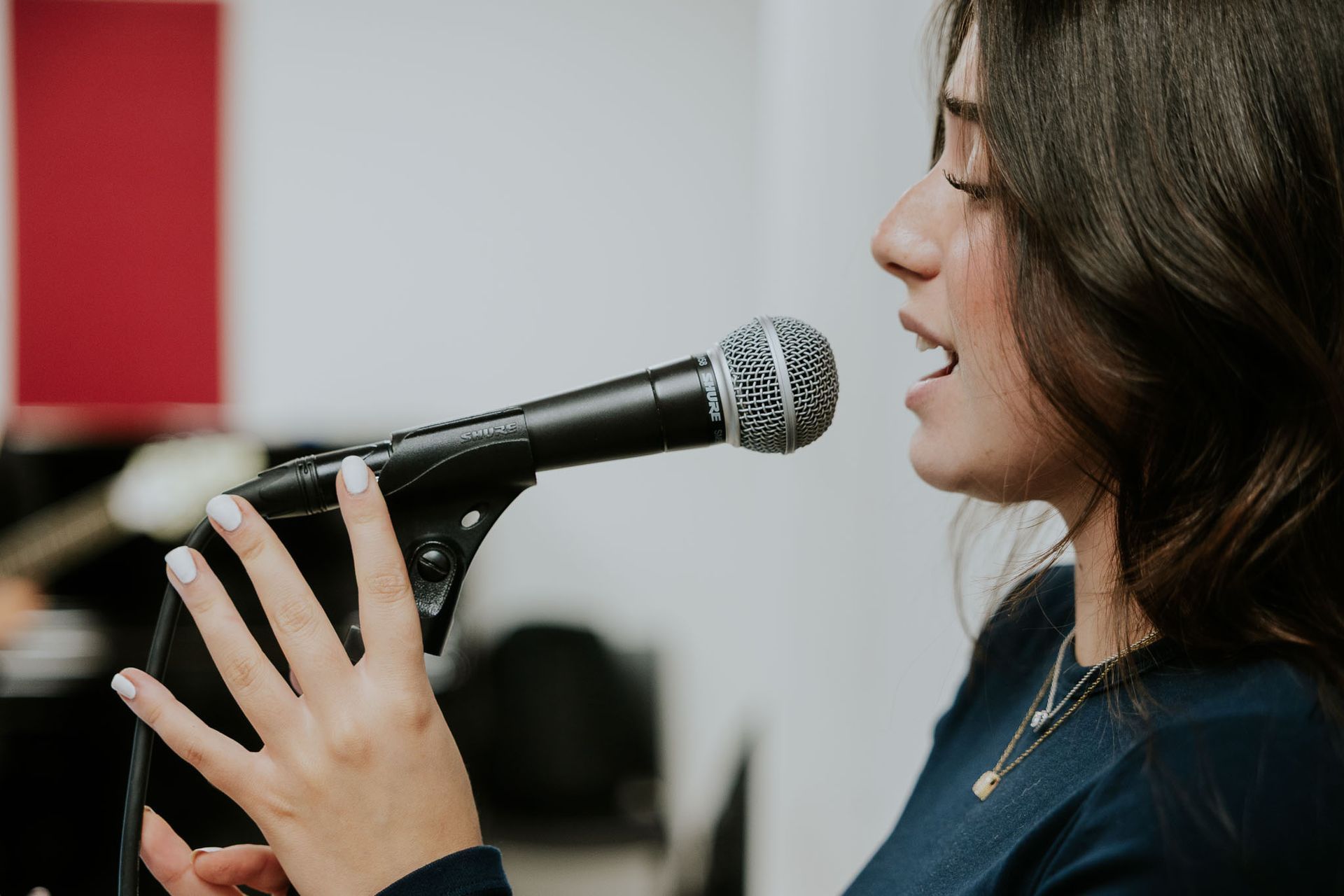 Donna che canta al microfono, in un interno. Indossa un top scuro, unghie bianche e una collana.