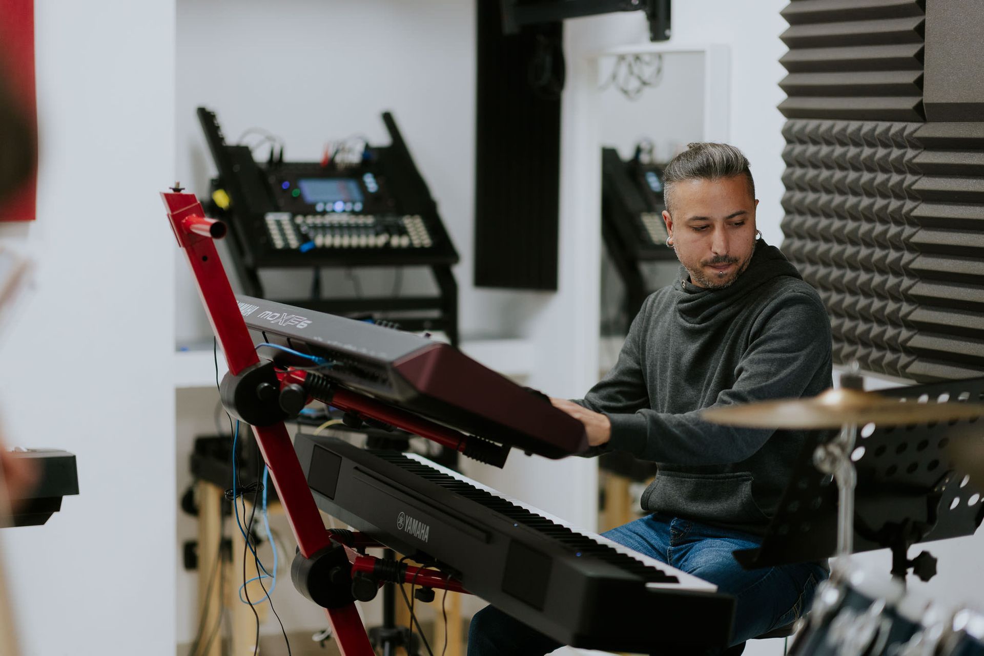 Uomo che suona la tastiera in uno studio musicale, espressione concentrata. Supporto rosso per tastiera, schiuma fonoassorbente visibile.