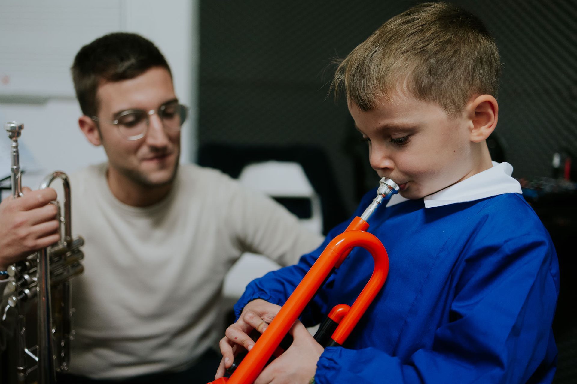 Un uomo aiuta un bambino a suonare la tromba arancione. Ambientazione in studio. Il bambino indossa un abito blu.