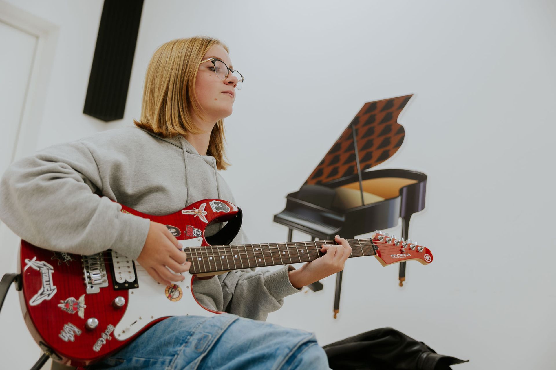 Persona che suona una chitarra elettrica rossa davanti a un murale raffigurante un pianoforte. Indossa una felpa grigia con cappuccio e occhiali.