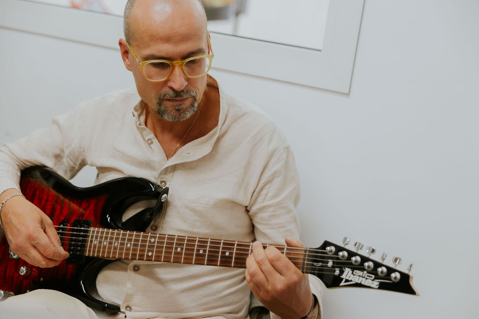 Un uomo con gli occhiali suona una chitarra elettrica rossa e nera in un ambiente chiuso, guardando attentamente verso il basso.