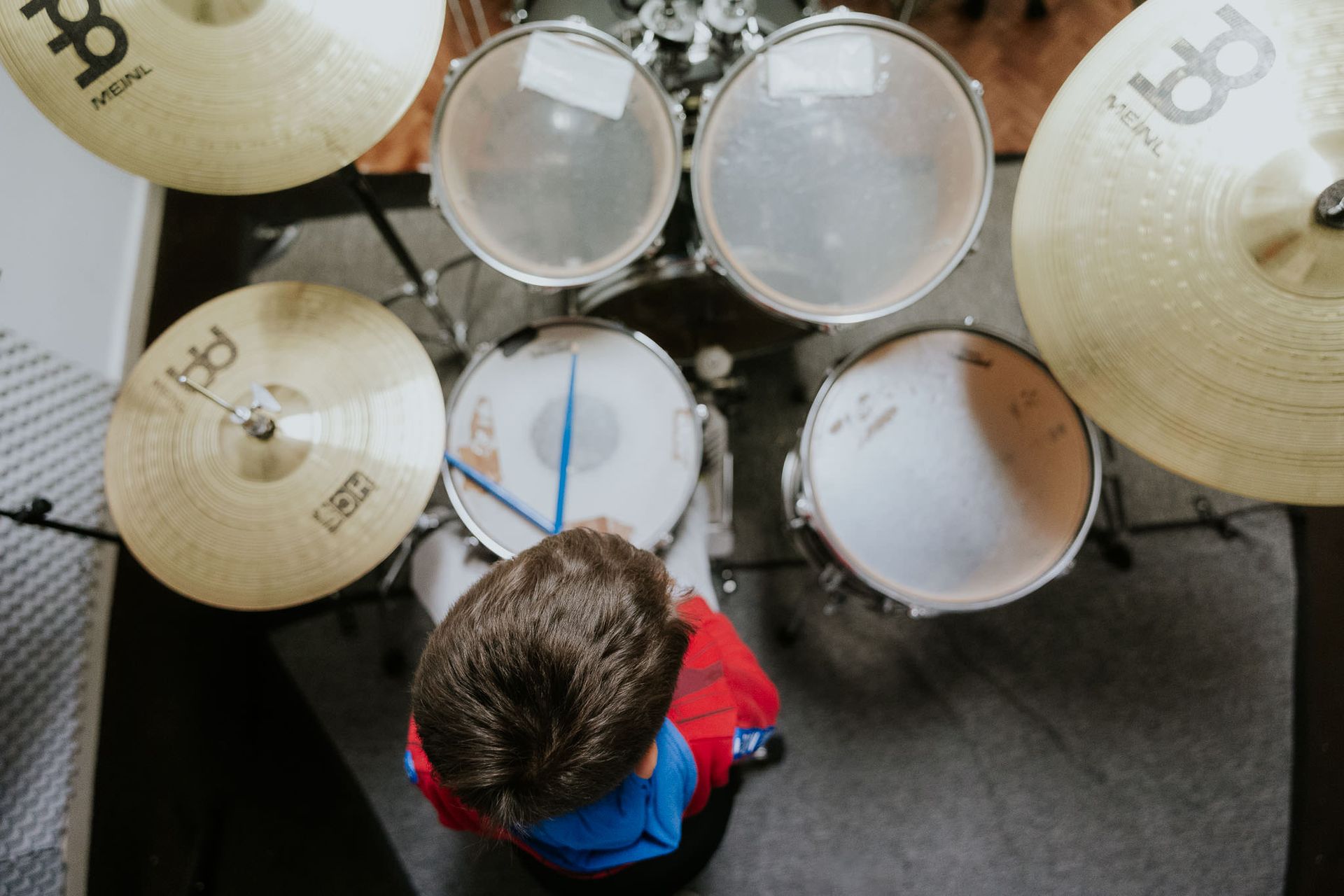 Vista dall'alto di una persona che suona una batteria con piatti. Indossa una maglietta rossa e usa bacchette blu.