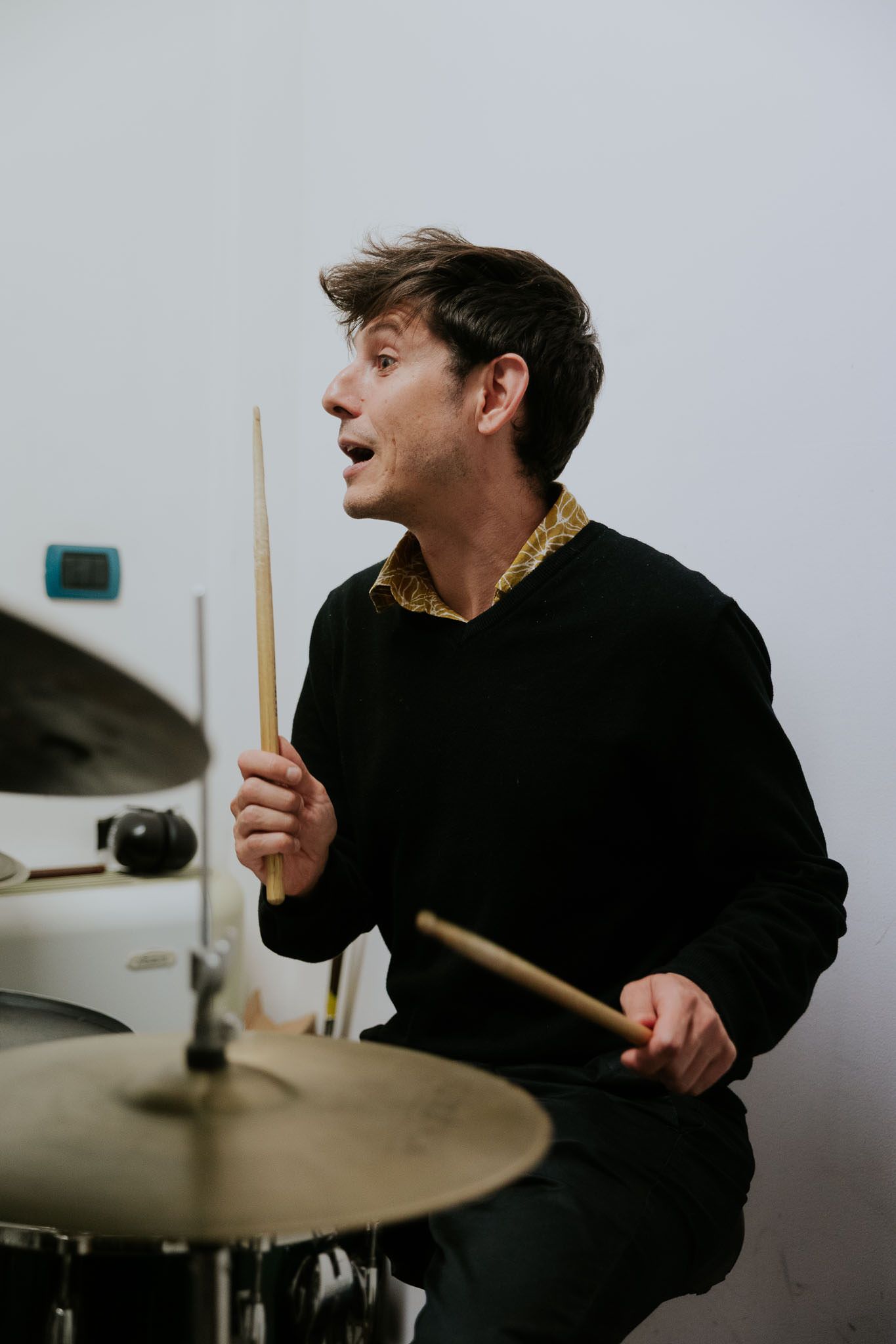 Un batterista suona, tiene in mano le bacchette e guarda in alto. Indossa un maglione nero, in una stanza bianca.