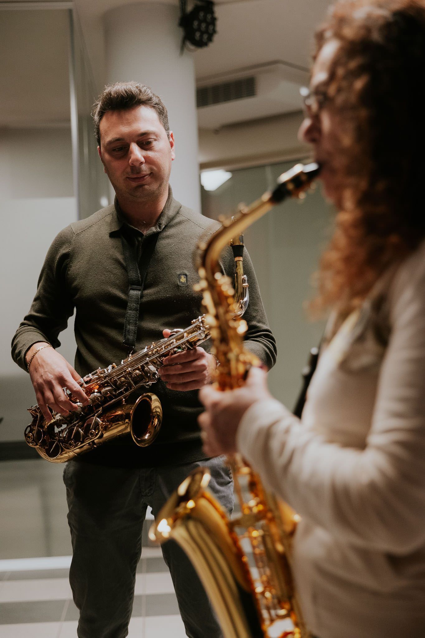 Due persone suonano il sassofono in una stanza. Uno guarda, l'altro suona. Strumenti dorati.