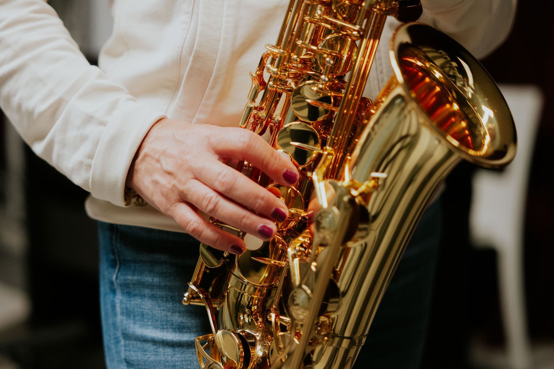 Persona che tiene in mano un sassofono dorato, primo piano. Indossa un maglione bianco e jeans, in un ambiente interno.