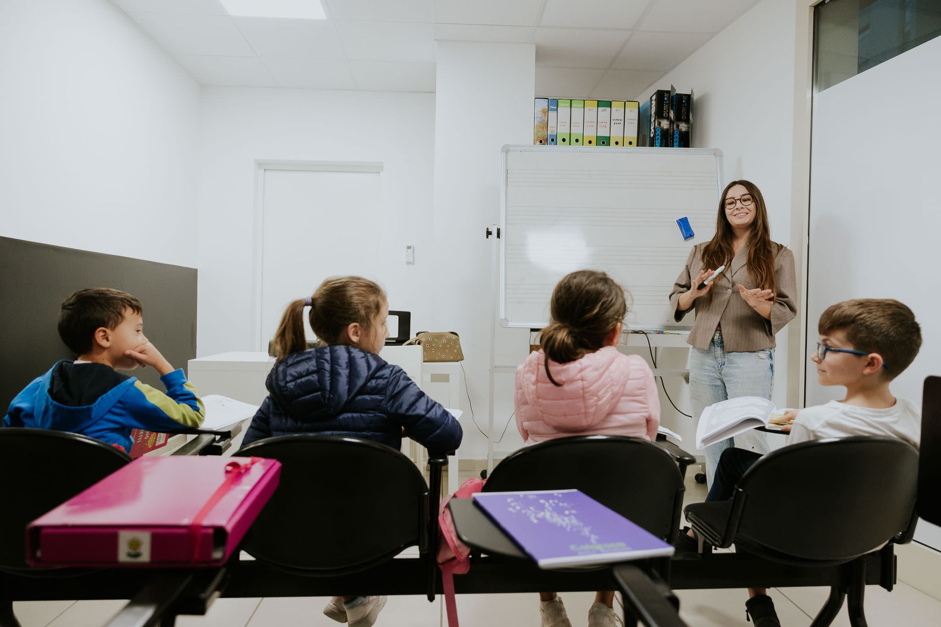 Insegnante in un'aula chiara, mentre insegna ai bambini. L'insegnante indica la lavagna, i bambini sono rivolti verso l'alto con i quaderni.