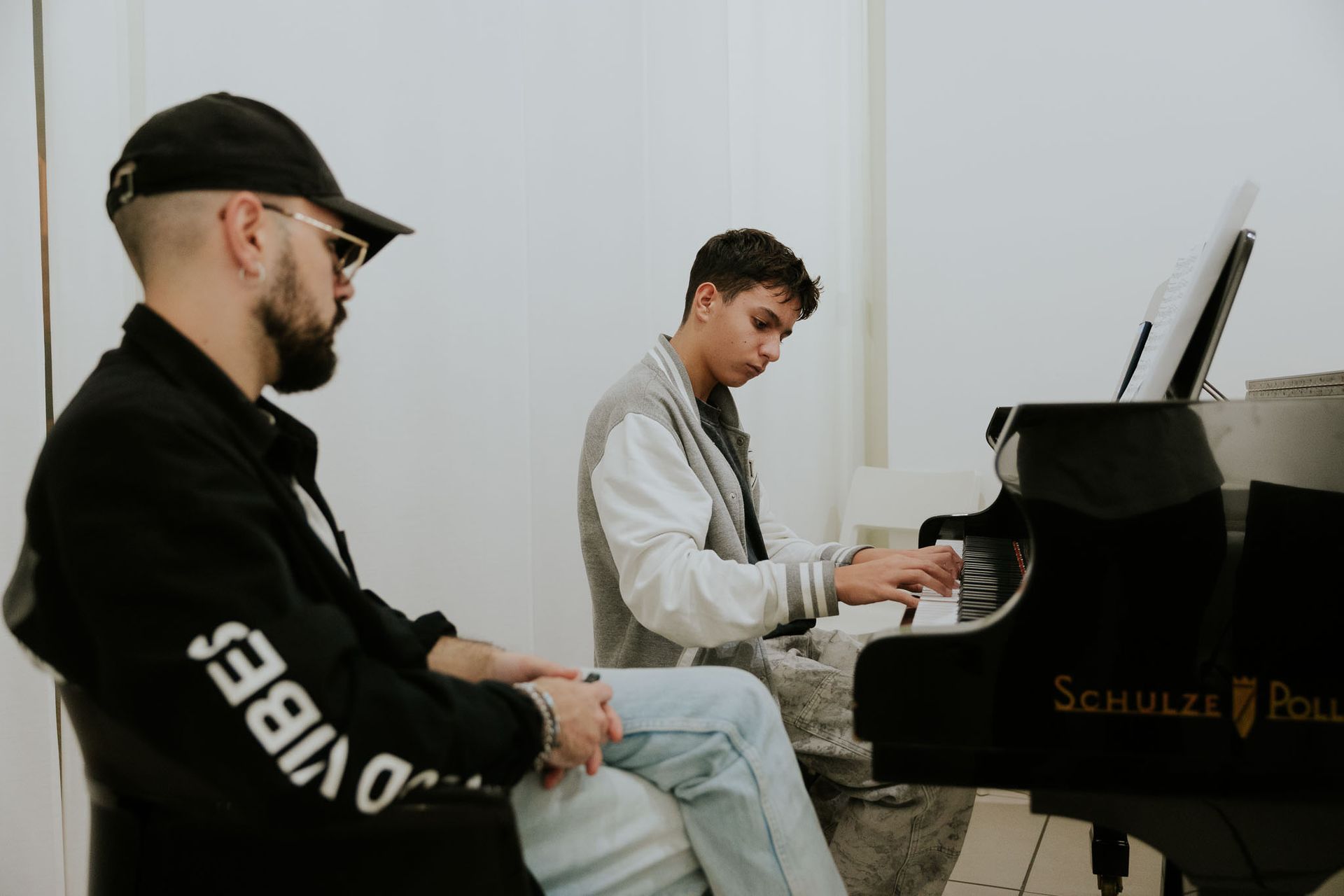 Un uomo guarda un'adolescente suonare un pianoforte nero. Ambientazione: stanza bianca.