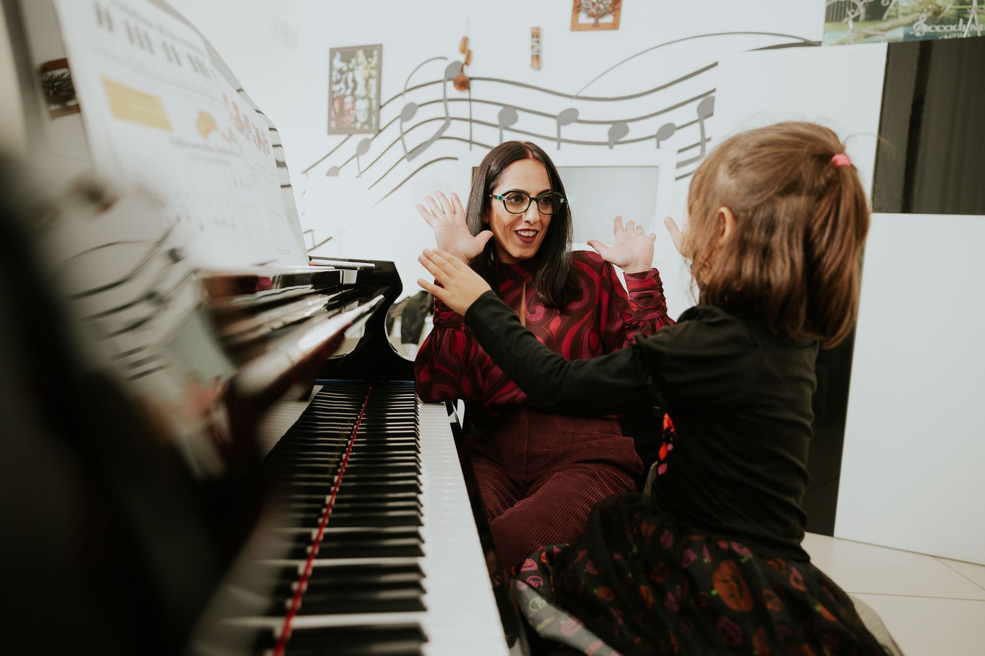 Donna e bambino con le mani alzate su un pianoforte, note musicali sul muro.