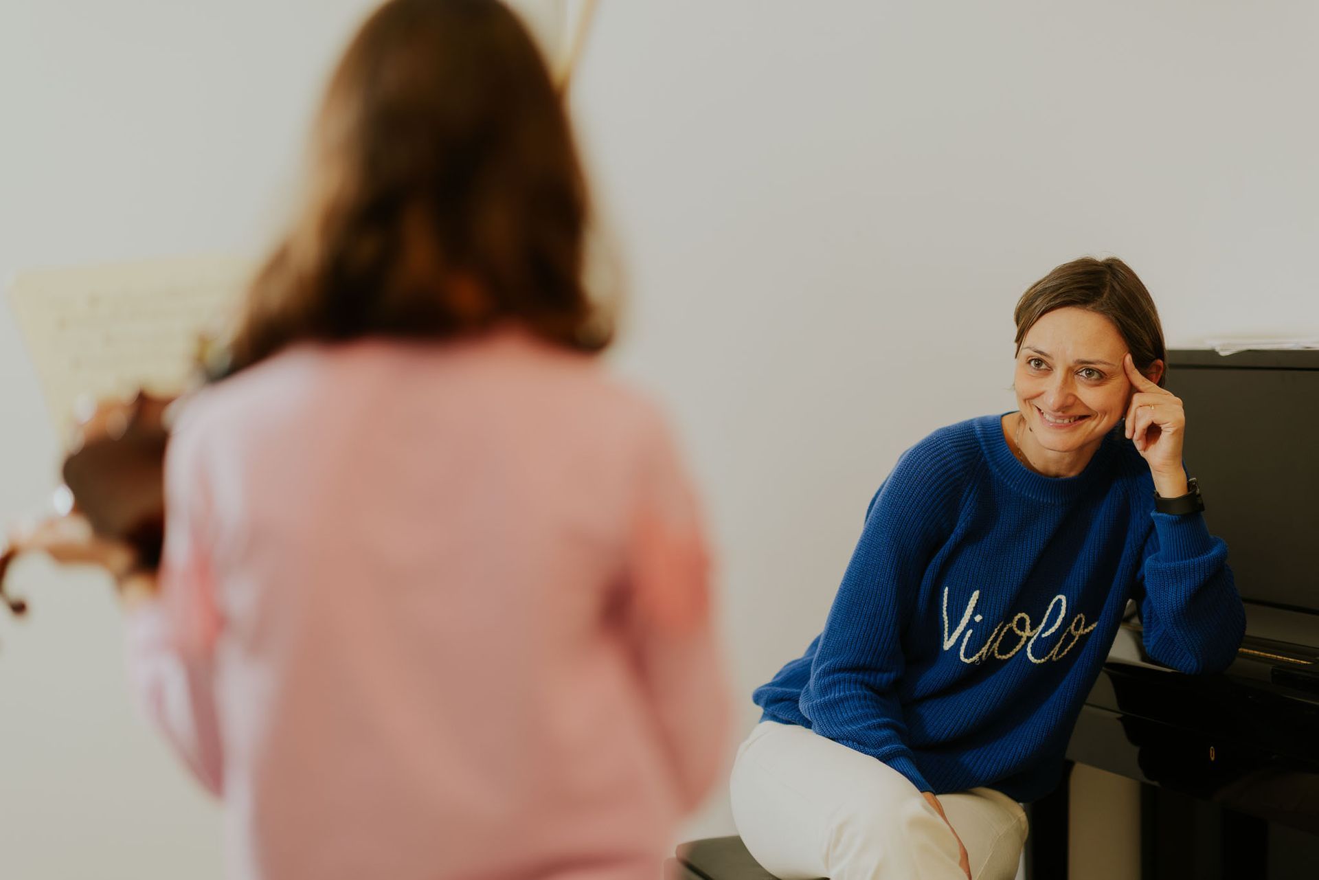 Una donna sorride a un bambino che suona il violino; la stanza è bianca e c'è un pianoforte.