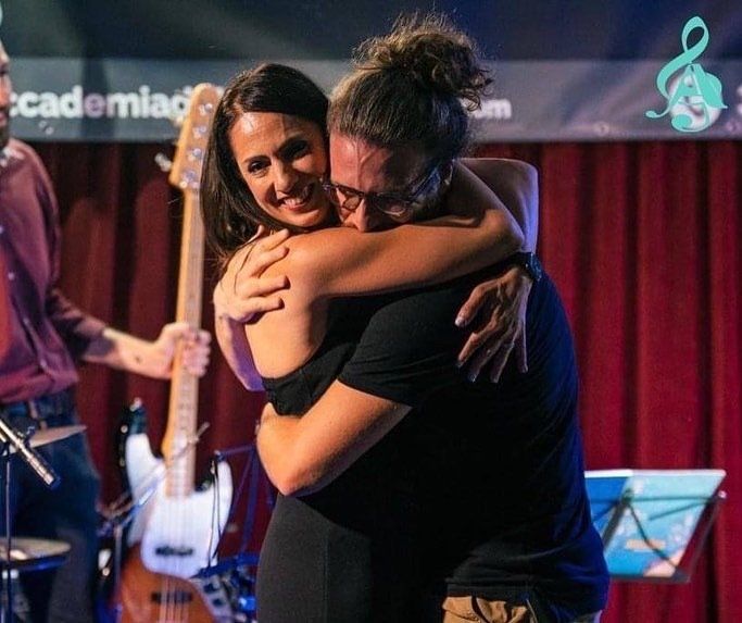 Due persone si abbracciano su un palco, sullo sfondo una band e delle tende rosse, una persona sulla sinistra tiene in mano un basso.