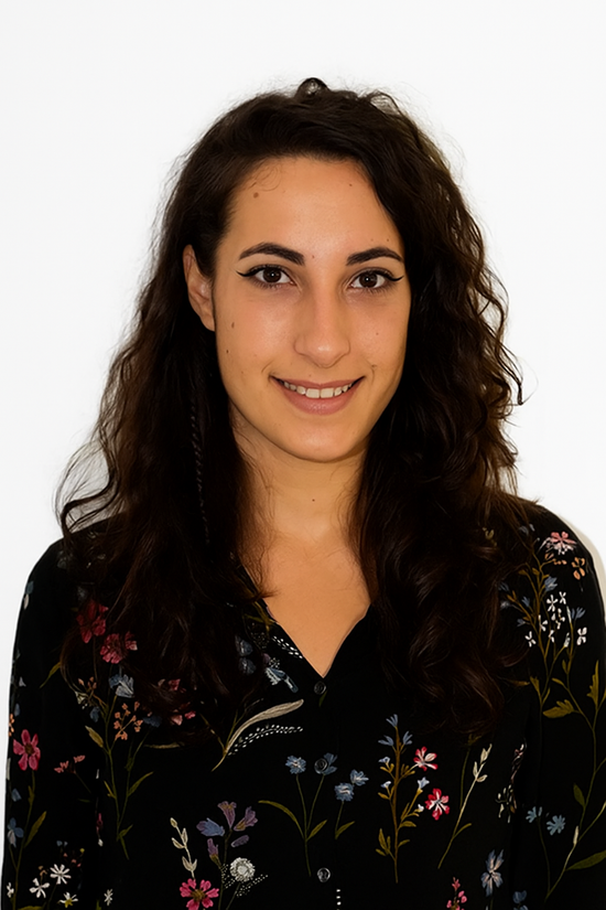 Una donna con i capelli scuri e ondulati sorride; indossa una camicia nera a fiori ed è in piedi su uno sfondo bianco.