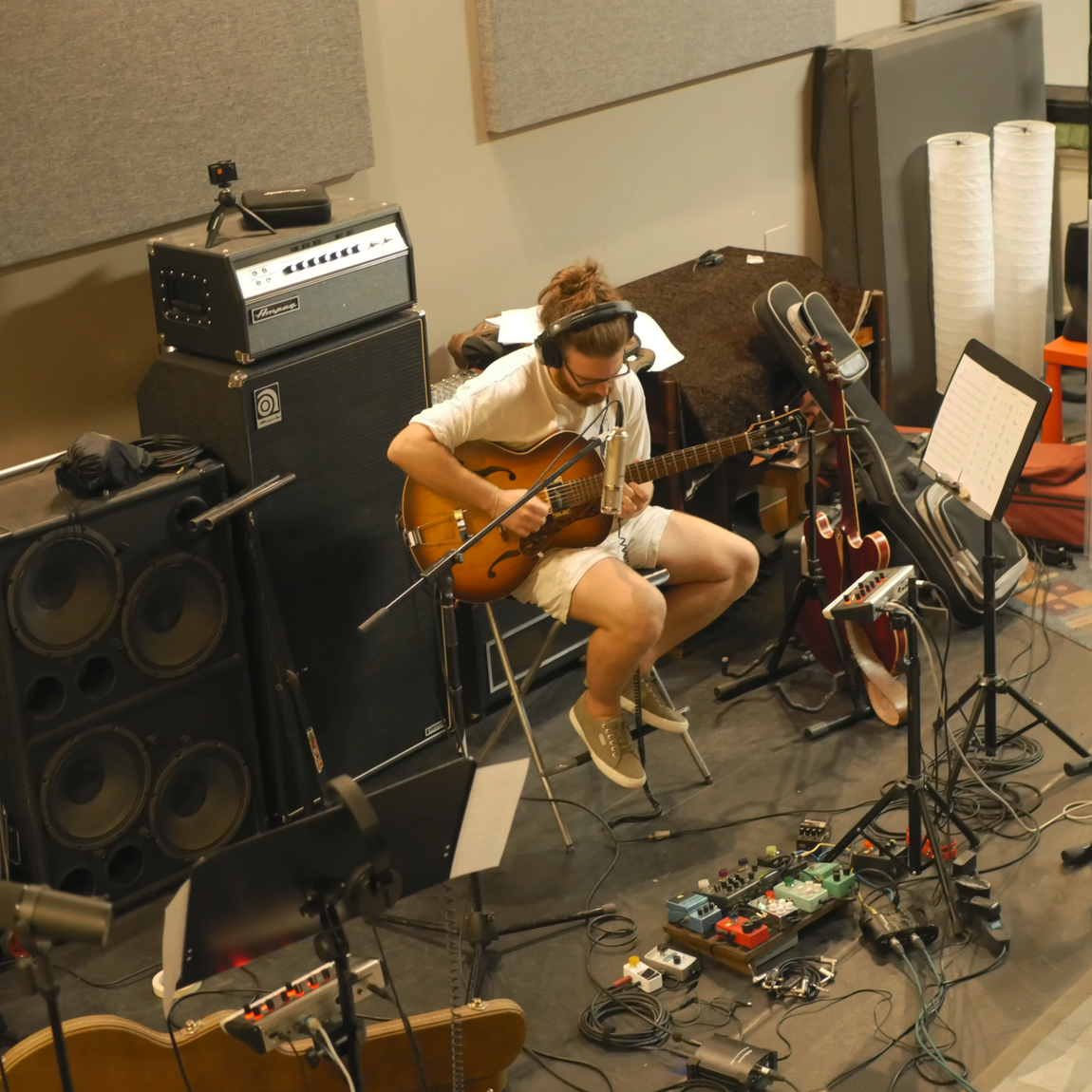 Una persona seduta suona una chitarra acustica in uno studio di registrazione con amplificatori, microfoni e pedali.