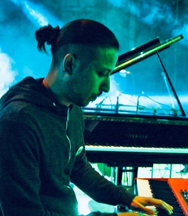 Uomo che suona il pianoforte, indossa una felpa scura con cappuccio. Ha uno chignon e i lati rasati. Palco illuminato con luce blu.