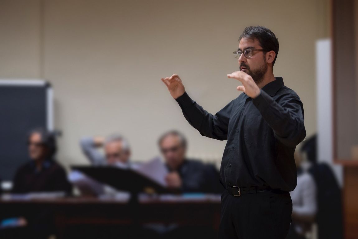 Uomo che dirige uno strumento musicale davanti al pubblico, con le mani alzate, occhiali e camicia nera.