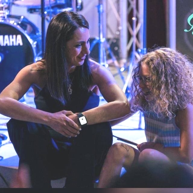 Una donna conforta una ragazza, entrambe sedute sul palco. Luci blu sul palco, batteria sullo sfondo.