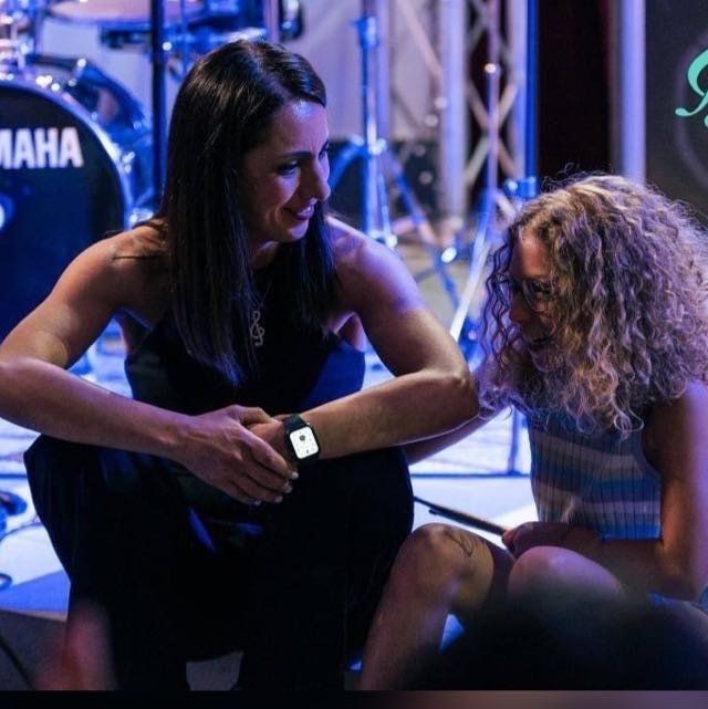 Due donne sedute sul palco, una che conforta l'altra. Palco con batteria e luci blu.